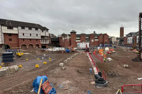 The former Riverside centre is demolished and levelled as part of a regeneration plan. There is a black and white building behind the building site, which has construction equipment and barriers on it.