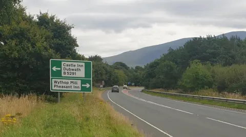 National Highways A single-carriageway stretch of the A66 with trees on either side.