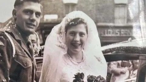 Mike Williams A vintage black-and-white wedding photograph. likely taken in the mid-20th century. Eric is dressed in a military uniform with a belted jacket and shoulder insignia. Mary is wearing traditional wedding attire, including a veil and a wedding gown, and is holding a large bouquet.