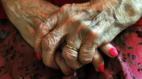 A stock image of the hands of an elderly lady overlapping and resting on her lap. Her nails are painted pink and she is wearing a wedding ring. She is wearing a pink skirt with a flowery pattern.