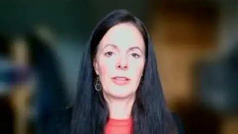 Sara appears on screen from a zoom recording. She has long dark hair and a red top under a dark jacket