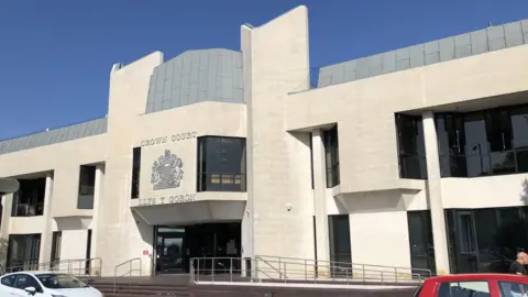 An exterior shot of Swansea Crown Court. There is a white car and a red car parked in front of the building. 