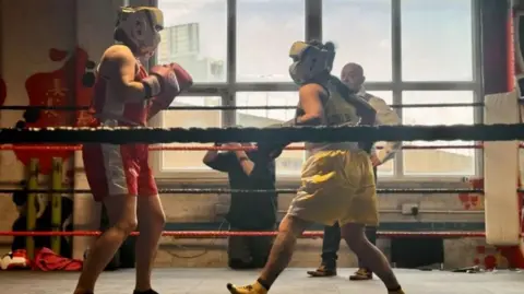 Two women, one wearing red boxing shorts and one wearing yellow boxing shorts. They are boxing each other in a boxing ring. 