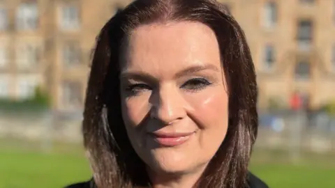 A headshot of Fiona Drouet. She has brown hair and is smiling. She is wearing dark eye makeup. A blurred building is visible in the background.