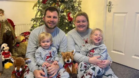 NHS Blood & Transplant Jodie, Amelia, Amelia's Dad and sister in matching Christmas pyjamas in front of their Christmas tree at home.