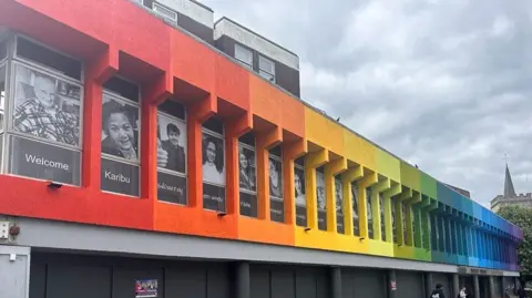 A rainbow coloured mural on at 70s-style building, with black and white pictures of different people on it saying, "Welcome", in different languages.
