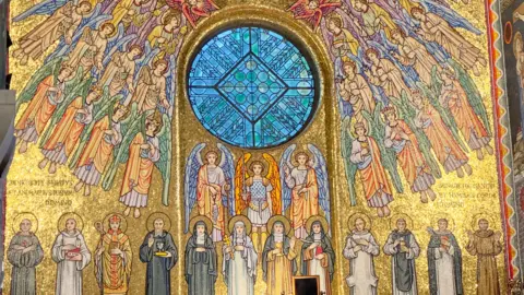 A predominately gold, detailed religious mosaic fills the image. Rows of winged angels surround a circular blue stained-glass window at the centre. A line of haloed saints stand below, all facing forward.