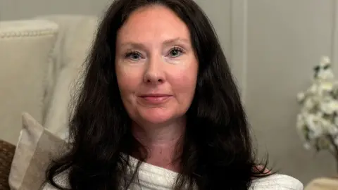 EuroMillions winner Ceri Roberts, looking at the camera. she is wearing a white dress, and a wall and flowers are in the background