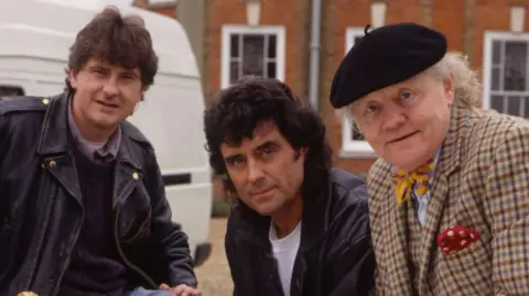 Actors Chris Jury, Ian McShane and Dudley Sutton leaning forward while looking directly into the camera. There is a white van behind them and a brick building