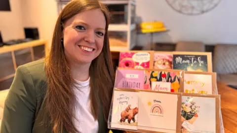 A smiling Andrea Pullen has dark red hair and is wearing a green jacket over a white T-shirt. Next to her on a desk is a stand displaying different birthday and Mother's Day cards.