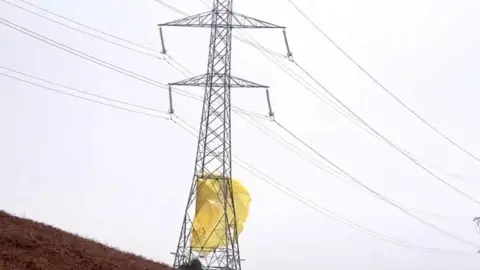 PA Media A large electricity pylon with yellow material resembling a parachute wrapped around the bottom of it.