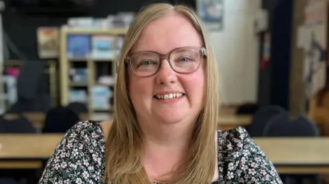 Kayleigh, mother of Oliver and Edward. She is a white woman with glasses and blonde straight hair. She is wearing a black dress with a pink floral print. She is sitting in a board game café.