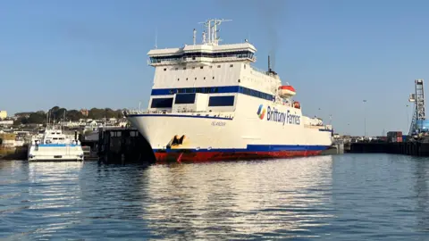 A Brittany Ferries vessel called Islander docked in Guernsey. The ferry is mostly white with some blue stripes. It is a sunny day.