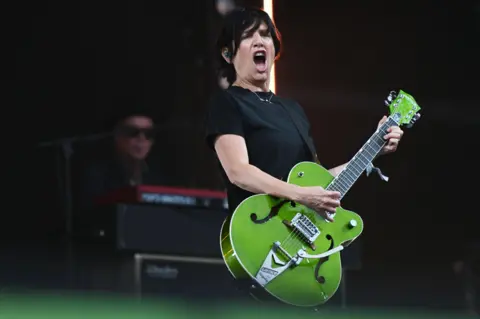 Texas star Sharleen Spiteri playing on the Pyramid Stage at Glastonbury. The singer is wearing a black-shirt and black jeans and is holding a green guitar 