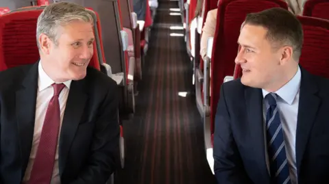 The PM sits across a train aisle from Wes Streeting, both smiling and looking at each other. The PM's tie is red, matching the seats and Streeting's is navy blue.