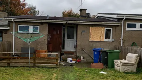 BBC A house with green grass in front of it. It is single storey with a white door and brown bricks. There is a clothes rail in the garden and a white armchair. There are three binds. A window on the right is boarded up.