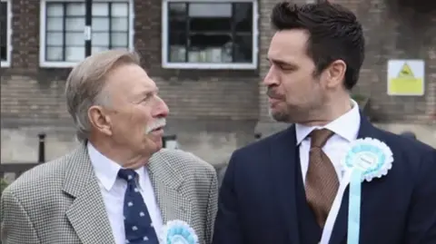 Alex Sibley A photo of two men wearing Reform Party rosettes, looking quizzically at each other outside an industrial brick building. The man on the left is older, with grey hair and a grey moustache, and wears a black and white houndstooth jacket over a white shirt and a navy tie. The man on the right has black hair in a duck tail style and wears a navy suit, a white shirt and a brown tie. 