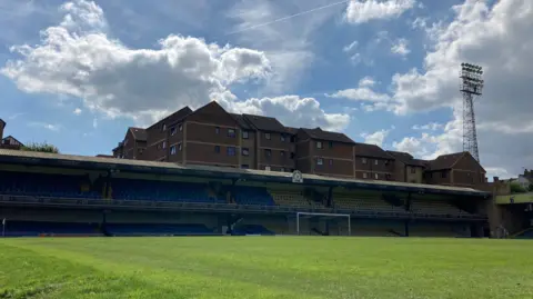 Tom Larsen Wright/BBC Inside Southend United FC's Roots Hall stadium. There is grass in front and beyond it is a stand. Behind it are flats and above them blue sky and white clouds.
