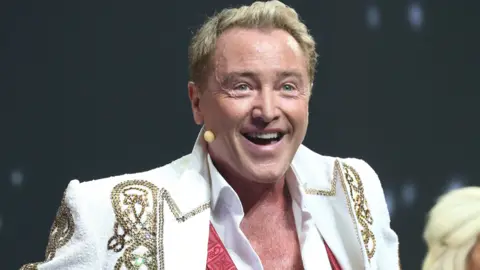 Michael Flatley is smiling as he performs on stage. He has short blonde hair. he is wearing a white blazer with gold trim and a white shirt which is unbuttoned down his chest.