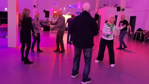 A room which is set up like a disco with pink lights and several older people dancing. 