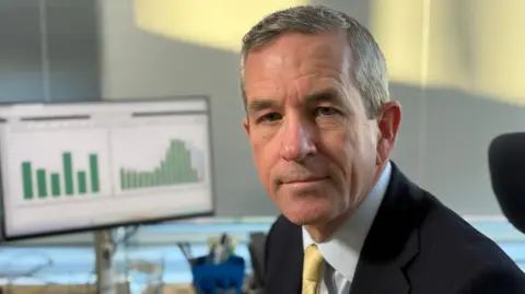 A man sitting at a desk turns to look at the camera. He has grey hair and is wearing a white shirt, pale yellow tie and dark suit jacket. In the background his monitor is displaying a graph showing a series of green columns on a white background. 