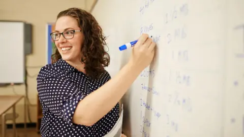 Getty Images Teacher with shoulder-length brown curly hair and wearing glasses stands with a blue marker writing on a whiteboard. Stock image.