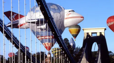 Bristol International Balloon Fiesta A hot air balloon in the shape of a virgin atlantic airplane flies near the iconic arch of clifton suspension bridge.