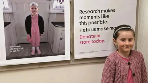 Faye in her pink cardigan smiles while posing in front of Cancer Research UK posters, which feature an image of her wearing a bandana and black and pink school uniform 