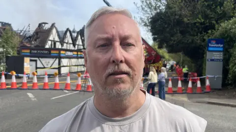 Richard Daniel/BBC A man with grey hair and a grey beard looks at the camera as he stands on a closed road in front of a nightclub where a fire has been. He wears an off-white top. People can be seen looking at the venue behind him. There is a cordon of cones in front of the venue.