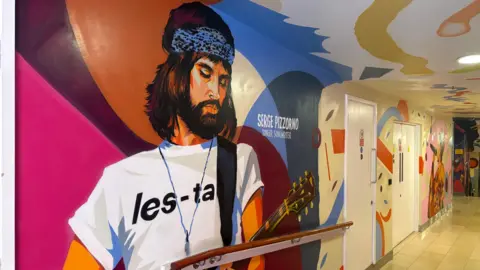 A street art painting of Serge Pizzorno, holding a guitar while wearing a white t-shirt with les-ta printed on the front.