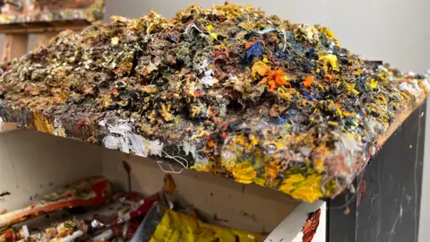 A mound of dried paint of all colours - underneath the first drawer is slightly open showing some of the tubes of paint it was squeezed from