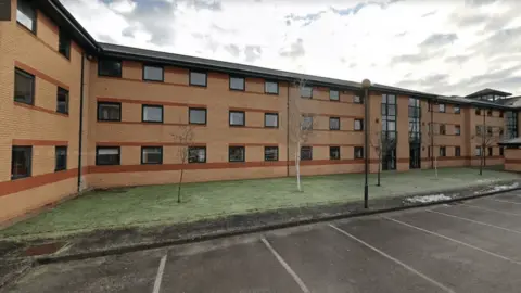 Staffordshire University halls of residence. The building is long, brick-built, and three storeys tall, with a grass area, trees and a car park in front. 