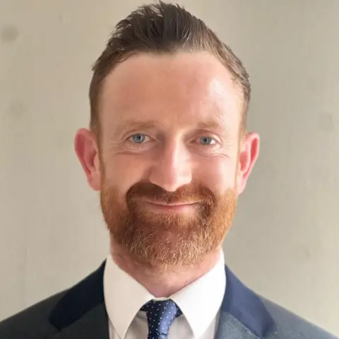 Scottish Family Party A man with shirt briwn hair and a ginger beard smiles at the camera. He is wearing a blue suit and tie