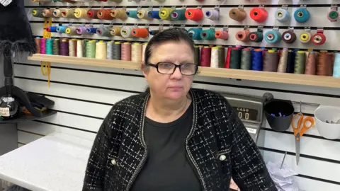 Tom Walker/BBC Boowy Latham, who has black hair and black framed glasses, speaks to the camera in front of a shelf holding spools of coloured thread.
