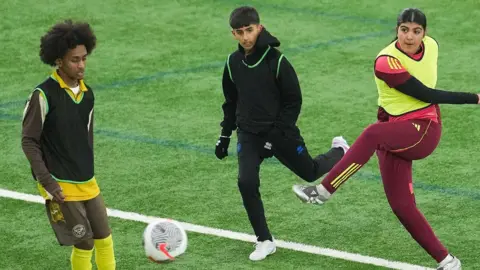 PFA Three young players taking part in the AIMS programme play on an indoor astroturf pitch. A young girl wearing a red tracksuit and yellow bib with black hair kicks the ball, and is next to two players, both with black hair. The player on the left is in a yellow and black kit and has a black vest. The second player wears a black tracksuit and is about to take a shot at the ball.