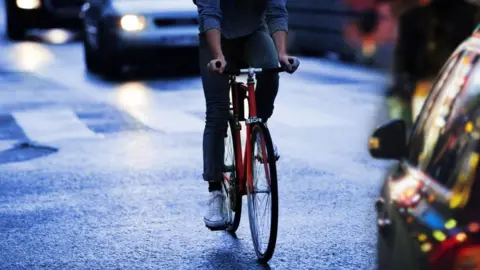 Getty Images A person riding a red-coloured bicycle along a road towards the camera and a car on the right. A car can be seen behind them with its headlights on.