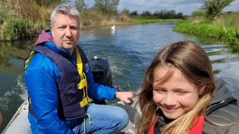 Family photo Emily and dad Andy in a ridged inflatable boat on the river. Dad is steering an outboard motor and is wearing a navy life jacket over a blue coat. The girl is wearing a red life jacket over a grey coat. 