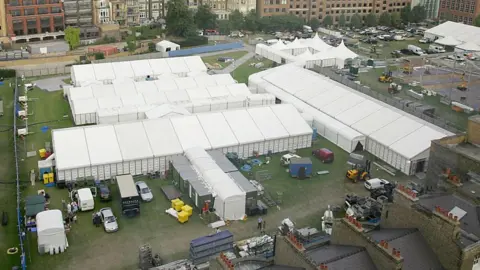 Getty Images Aerial view of the temporary mortuary, featuring numerous long white tents arranged in rows on a grassy field. The site is surrounded by vehicles and equipment, such as bright yellow bins.