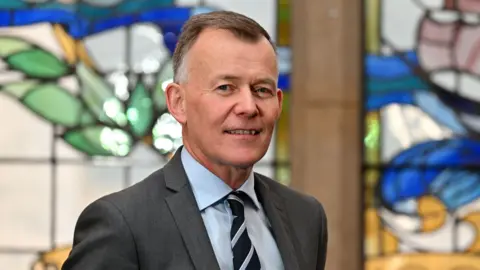 PA Media A man is wearing a grey blazer, blue shirt and striped tie. He is standing in front of stained glass windows. He has short brown hair. 