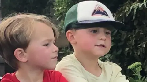 Family Handout Louis wearing a red top standing next to George who is wearing a yellow t-shirt and a blue, green and white cap