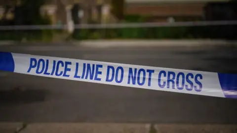 A police corden in white and blue that reads "police line do not cross". The background is a blurred pavement. 