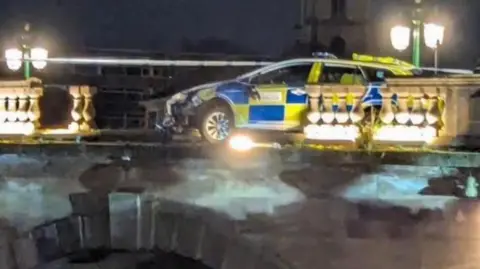 A police car that has driven through a balustrade on a bridge. The car is partially overhanging the bridge edge. It is night and the scene is lit by lampposts.
