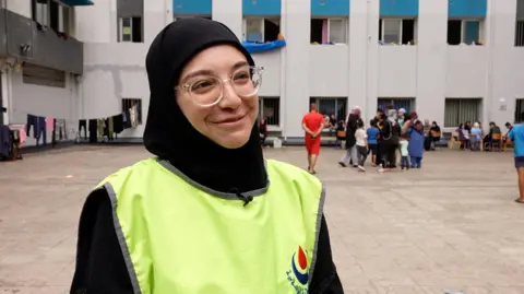 Batoul Hammoud stands in the schoolyard wearing a high-vis jacket and round glasses.