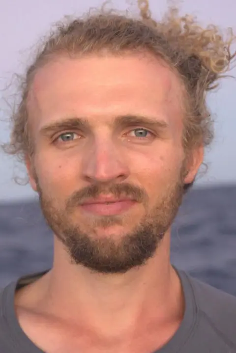 The MacLean brothers Lachlan has blonde wavy hair and a short beard. He is wearing a grey t-shirt and has the sea behind him.