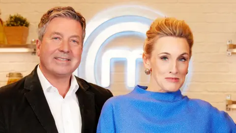 BBC John Torode and Grace Dent pose while looking at the camera in front of the Masterchef logo in a publicity still for Masterchef