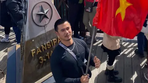 Ike is holding the Vietnamese flag on a balcony. Other tourists are surround Ike - they are taking photos of the view. Ike is wearing a black top with a necklace. Ike had a pyramid shaped statue behind him. The statue has a star on it. 