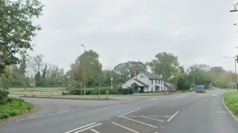 A junction on the A134 on Lynn Road. In the distance past the turning is a white property and a blue van driving away.