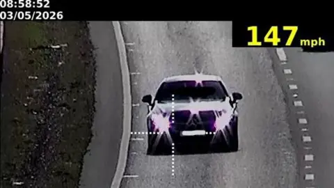 A black and white CCTV-style image of a Mercedes with the light reflecting off its lights and bonnet driving on the A14.