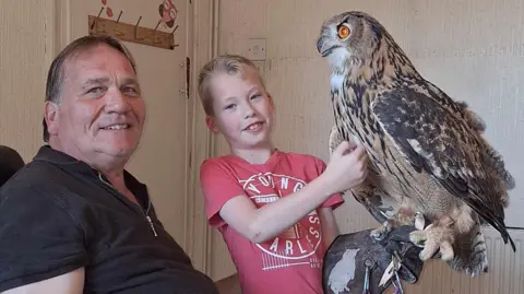 A boy with blonde hair in a red T-shirt holds an owl on a gloved hand in a living room. A man in a black top sits next to him. Both are looking at the camera.