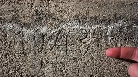 Scratches in a grey concrete wall which say "1943". There is a finger pointing at it from the bottom right hand corner.